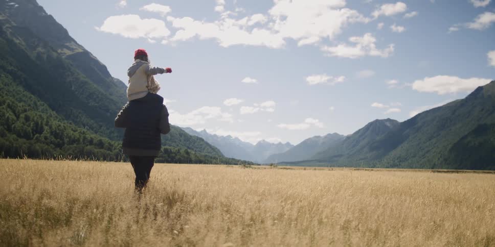 A child rides on a parents shoulders and excitedly points up to the blue sky and hills