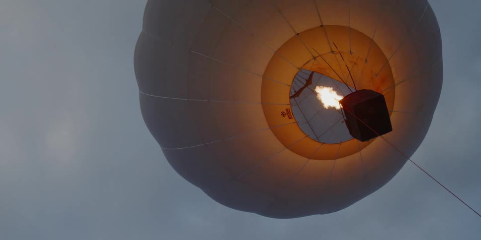 Flames burst into a hot air balloon against a grayish blue sky