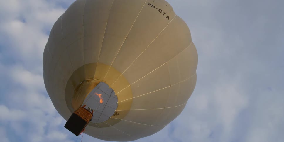 Flames burst from the burner in a floating hot air balloon against a blue sky with white clouds