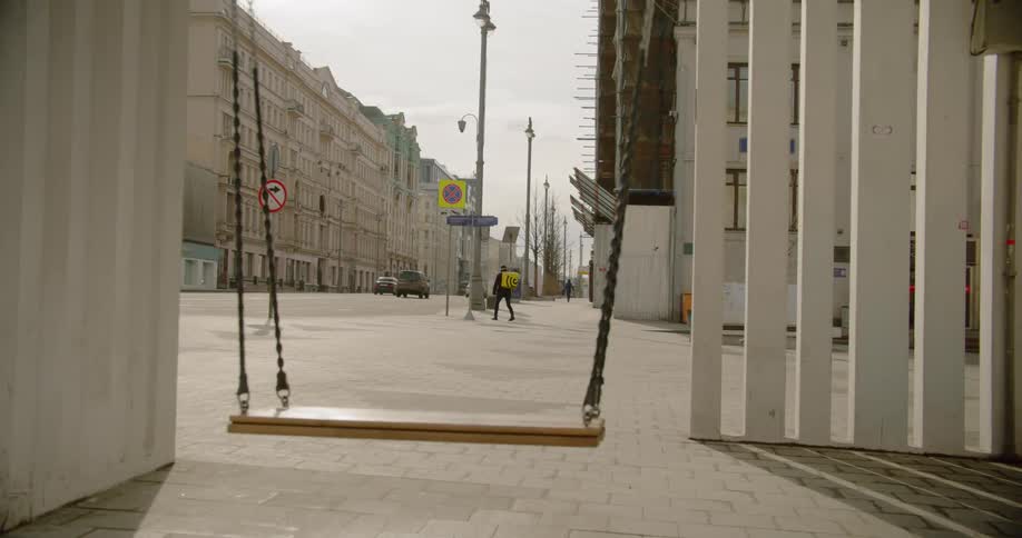 A swing sits empty in a city as cars drive by and two pedestrians walk on the quite sidewalk