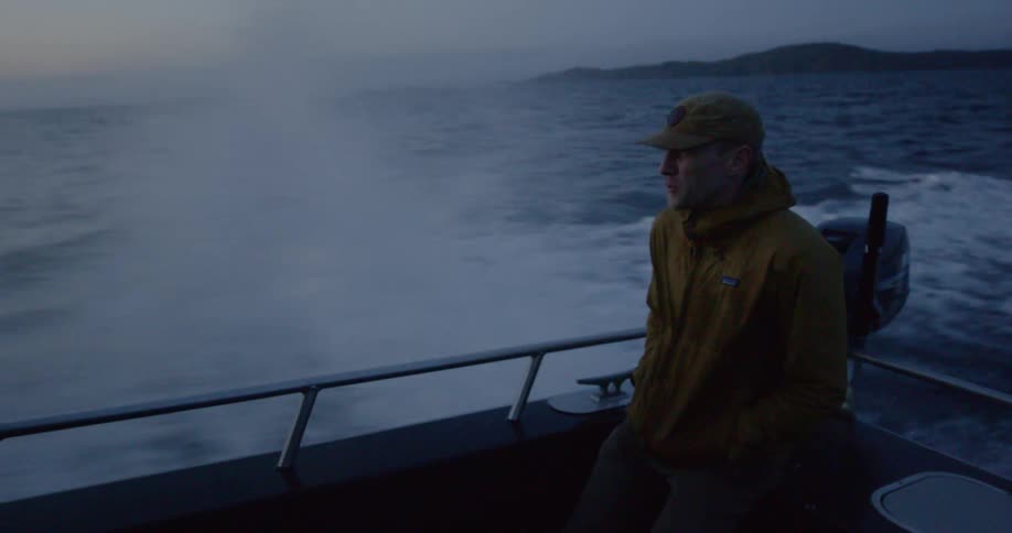A man leans with hands in pockets against the back rail of a speeding boat as waves splash