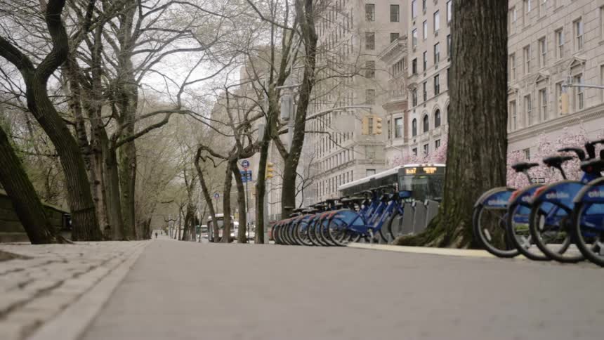 A public city bus travels down the street beside large buildings and a row of bicycles