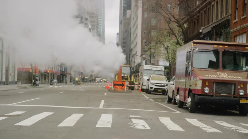 A city street with vehicles buildings and steam coming out of the sewer system