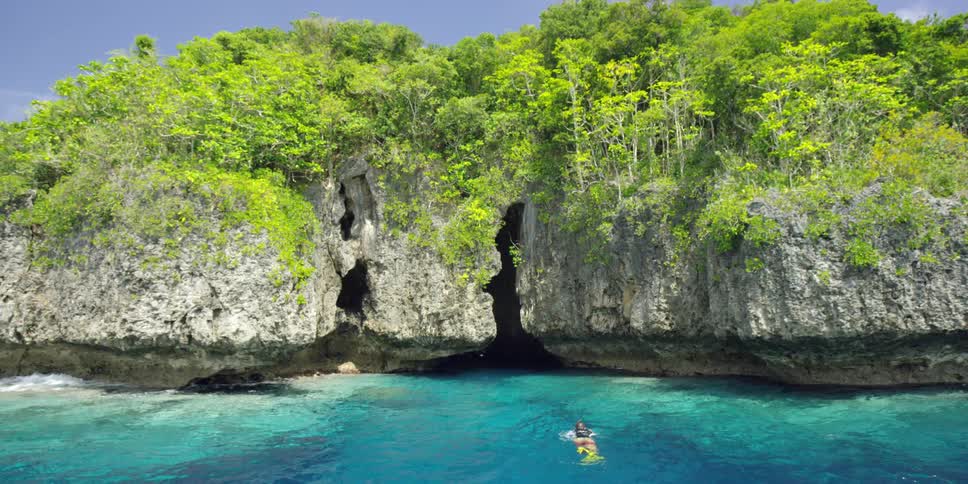 A man swimming towards a cave set in the side of a very steep cliff