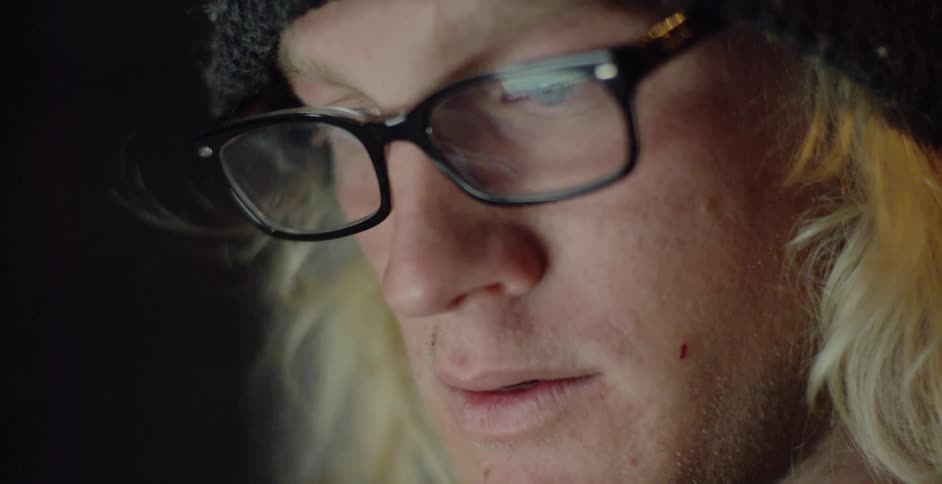Blonde haired man wearing glasses and a knit cap studies monitor screen