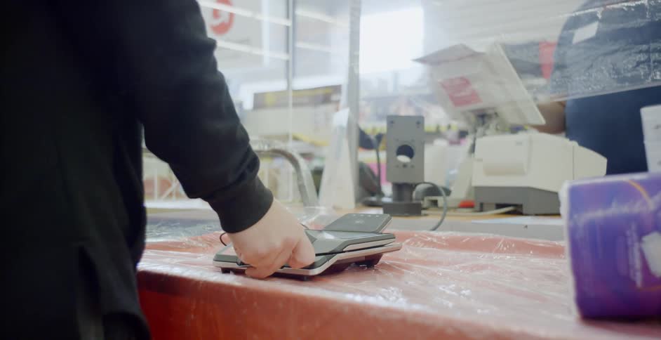 A person putting a credit card in a credit card reader with a cashier behind the counter with mask and gloves