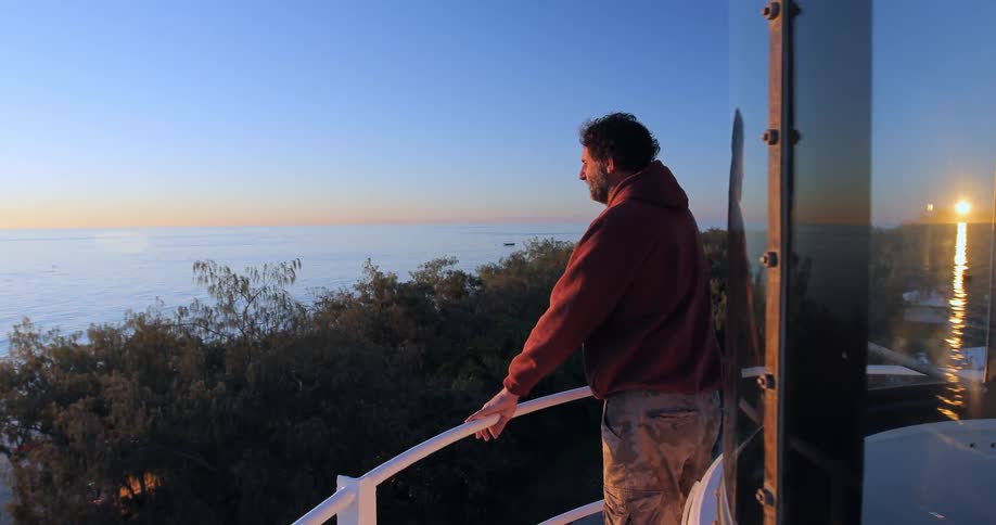 A man is standing on the balcony of a light tower by the water