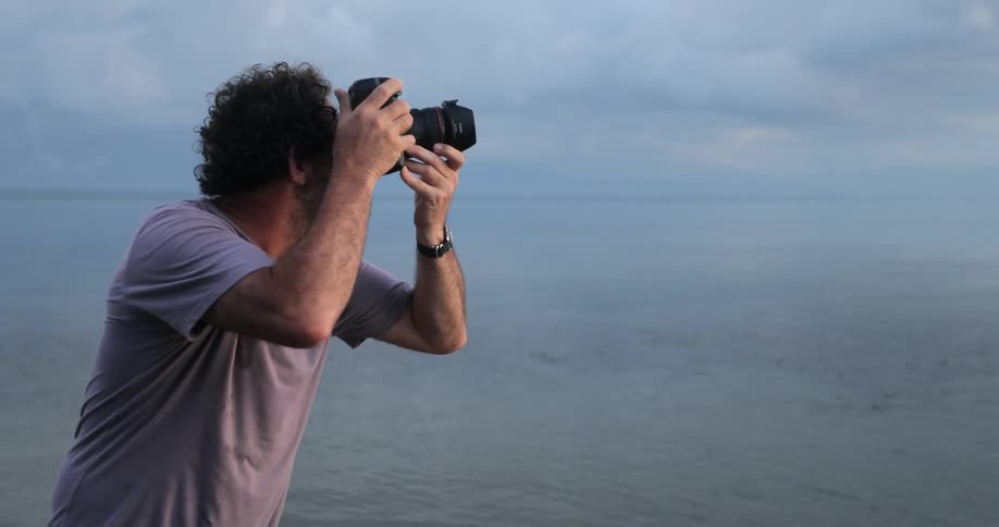 A man is standing on the beach taking photographs of the sun setting