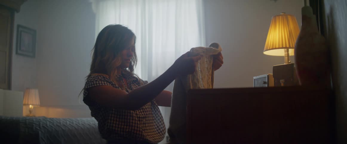 A woman kneeling in front of a dresser holds up a large sweater then hugs it and starts to cry