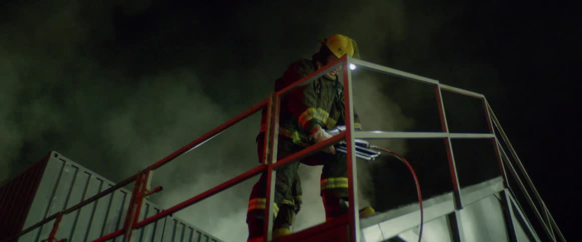 A firefighter positions a tool on a roof while smoke billows behind him at night