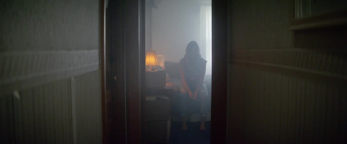 A person sitting on a bed in a bedroom at the end of a dark hallway
