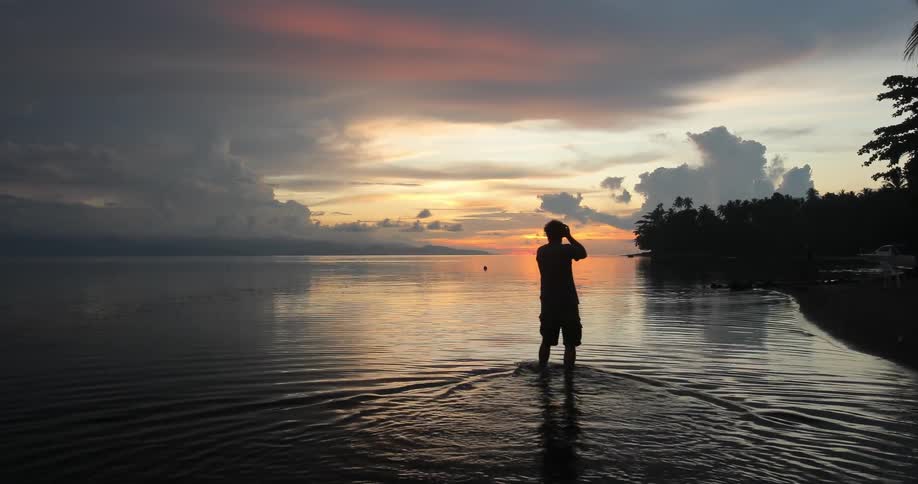 A man wading in calf-deep water photographing the sunset on a cloudy evening