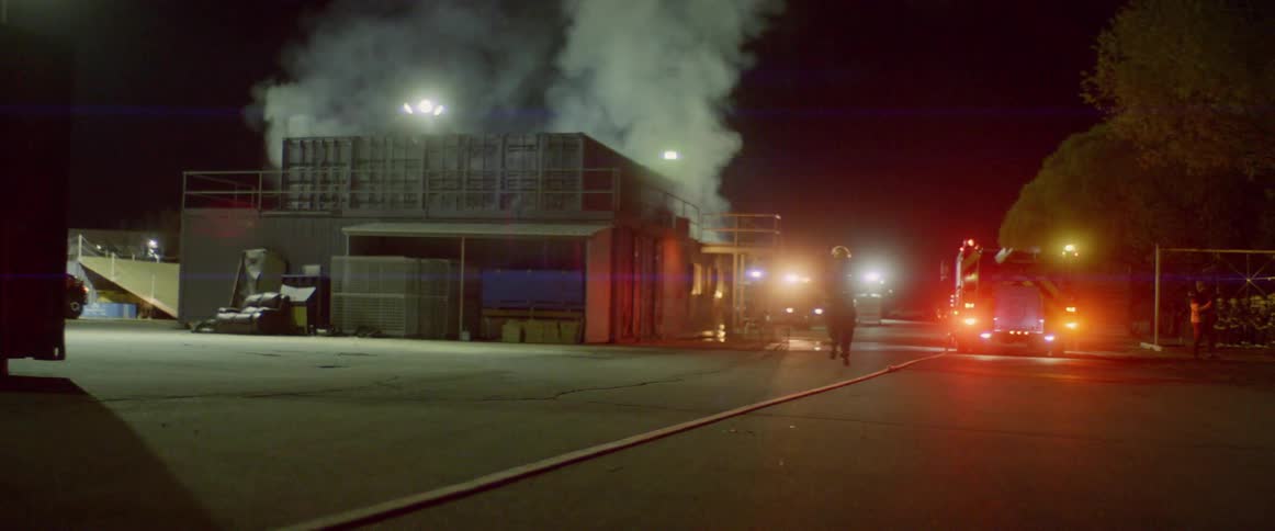 Firefighter carries a firehose towards a burning building as lights flash on the firetruck
