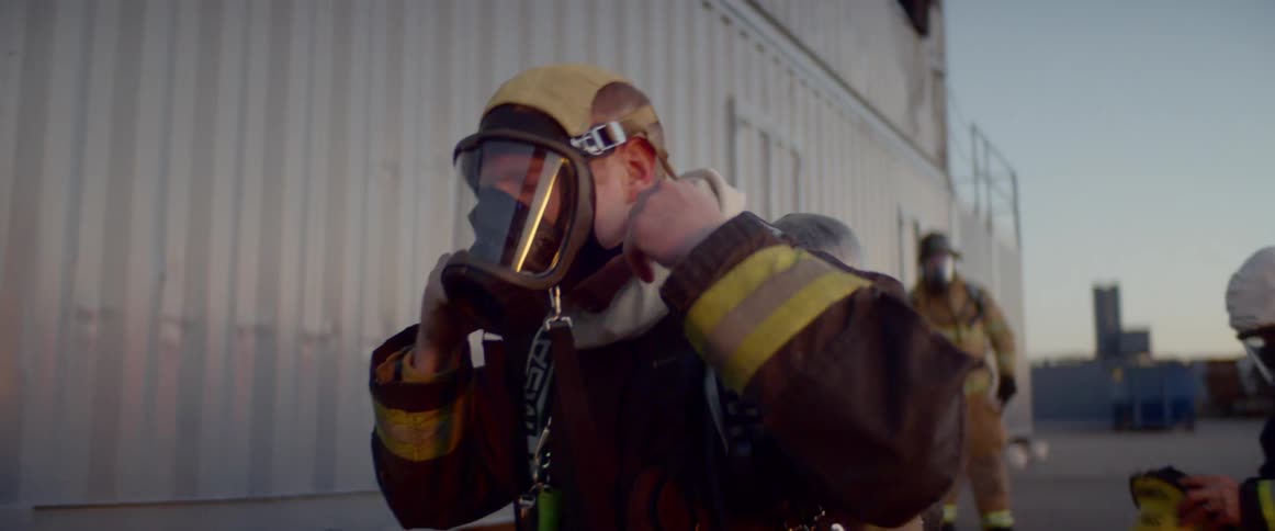 Three firefighters stand outside a building while two of them finish putting on their gear
