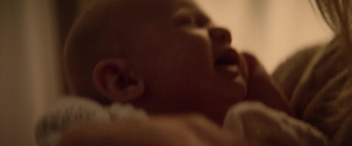 A baby cries with an open mouth while his mom tries to comfort him by rocking and shaking