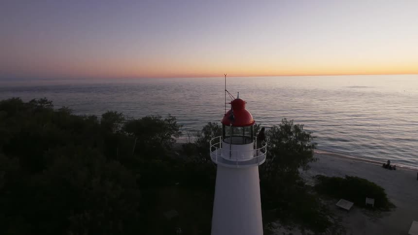 Drone footage of someone standing on a lighthouse deck as other people hang around on the beach below at sunset