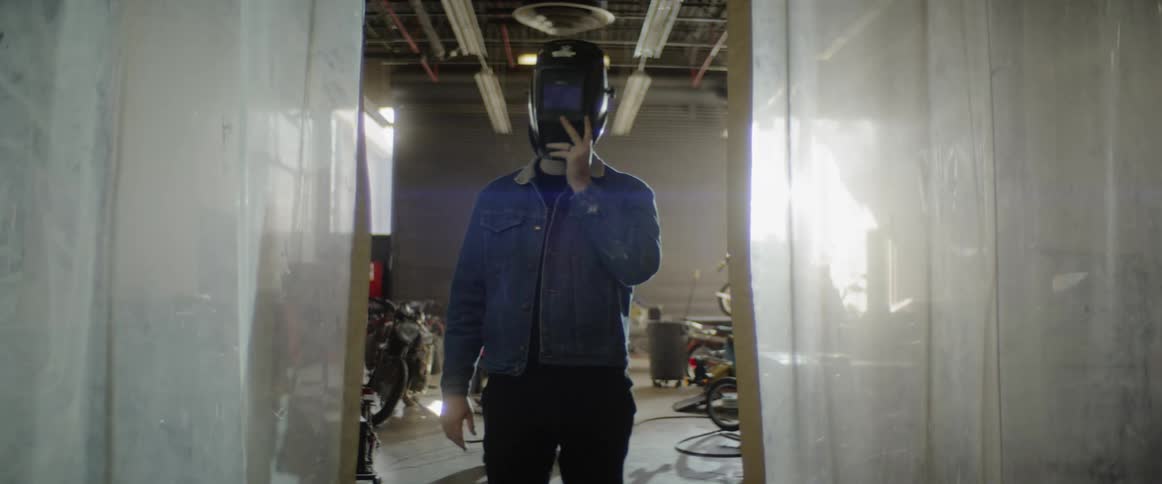 A man standing in between hanging plastic dividers takes off a welding helmet and looks at it