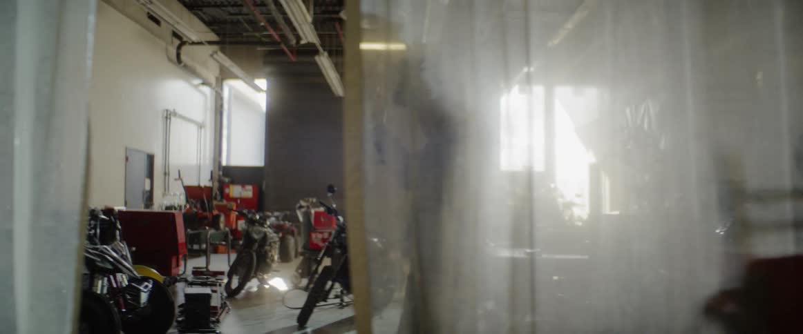 A man walks past hanging plastic divider in a motorcycle repair shop and puts on a welding helmet