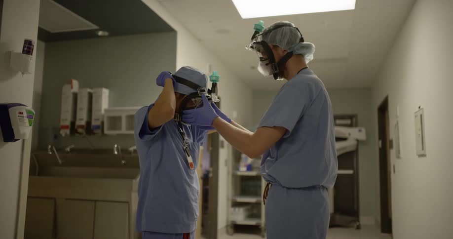 One medical professional helps another secure a face mask shield while in a hospital hallway