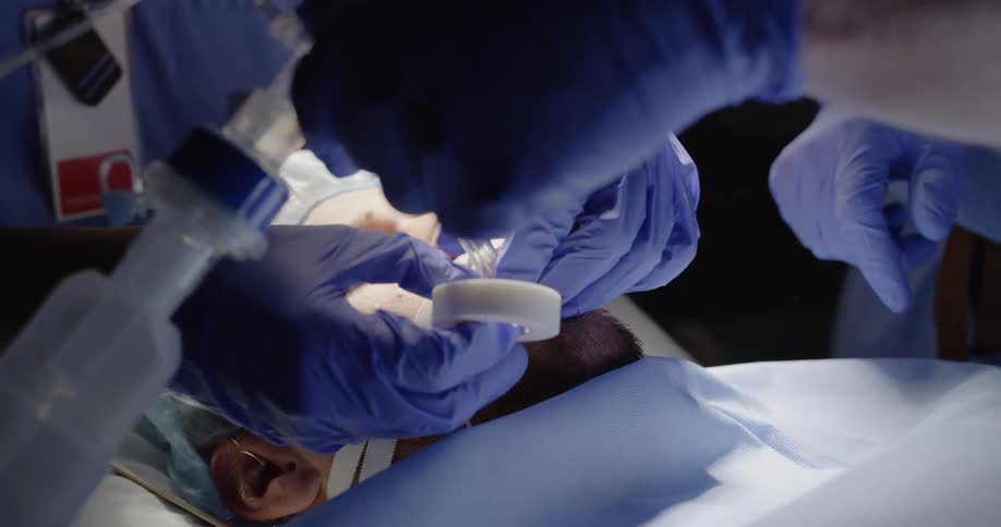 Healthcare workers wearing blue protective gloves wrap medical tape around an intubation tube while the male patient remains unconscious