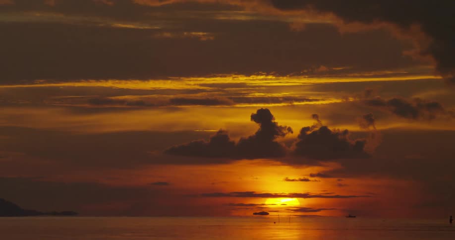 The sun is large and glowing orange as it sets, lighting up the sky behind dark clouds above the ocean