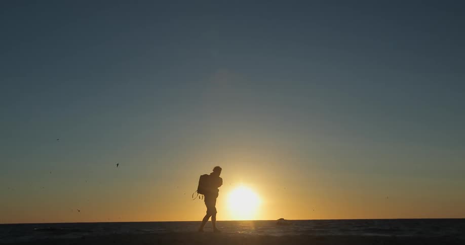 A person picks up their bag and walks across the beach at sunset
