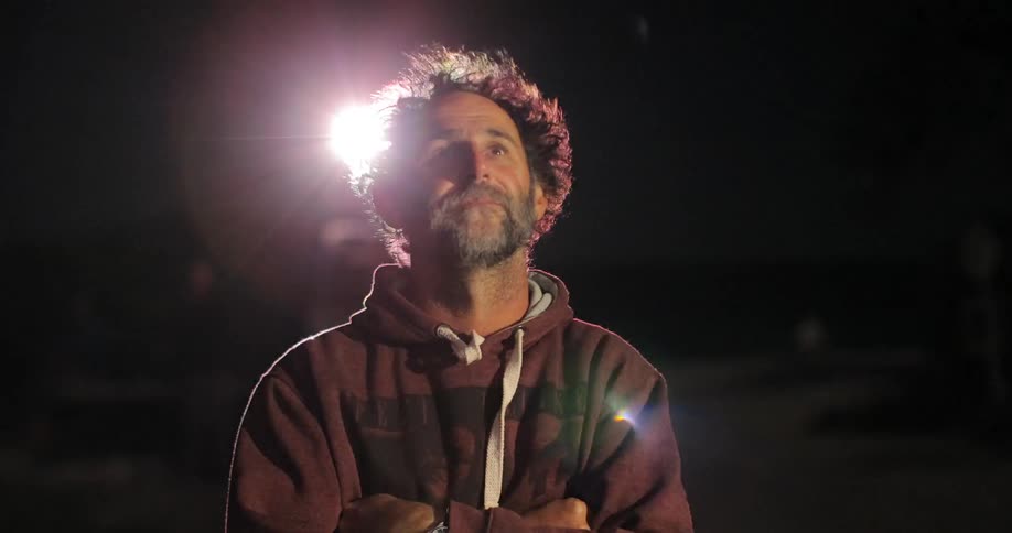 A man stands outside alone in the dark with a bright light shining behind him