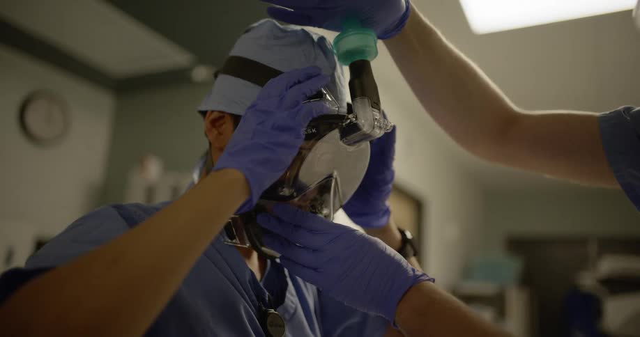A medical professional helping another put on a plastic gas mask