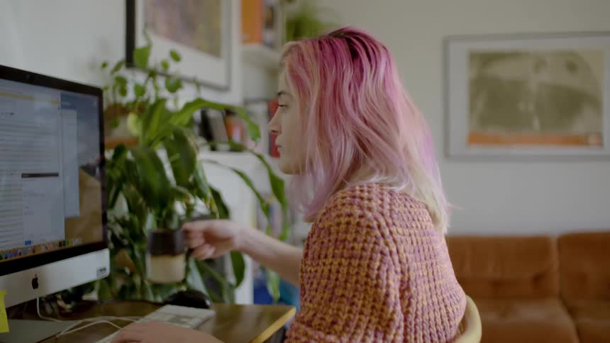 A woman types on a computer at a desk in her living room and stops to drink coffee