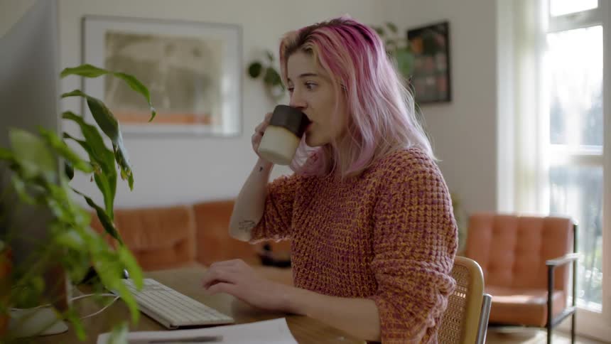 A woman sits at her computer in her living room pausing to drink some coffee