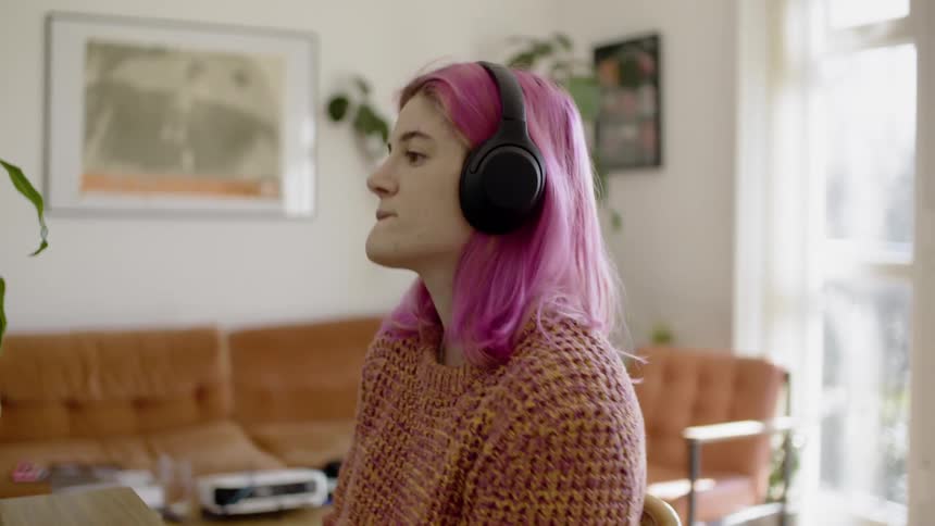 Pink haired woman sitting at the desk and wearing headphones takes a note and listens to something through headphones