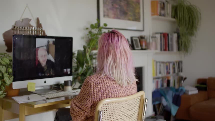 Pink haired woman sitting at the desk makes a video call on the desktop in her room during daylight