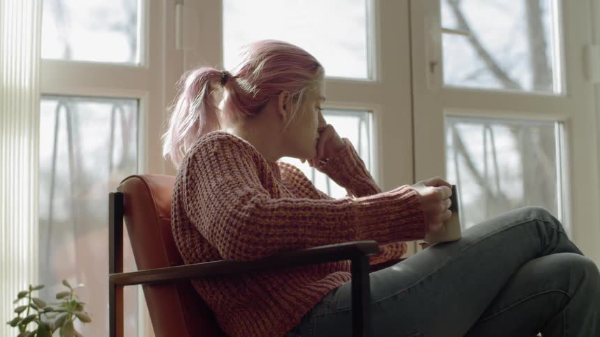 Female sits in a chair next to a window holding a coffee mug on her lap with her face resting on her closed hand looking depressed