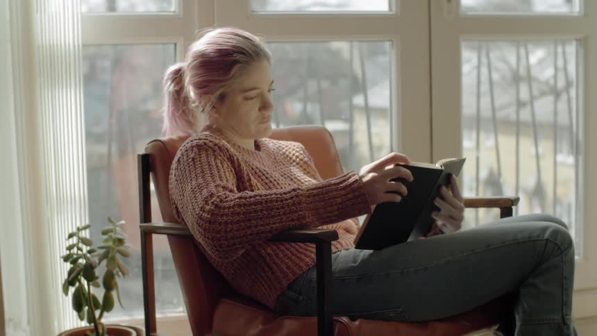 A woman reading a book while relaxing in a chair near a set of windows