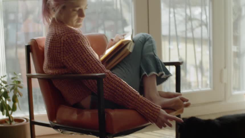 Pink haired female sits on a chair in her living room by her cat
