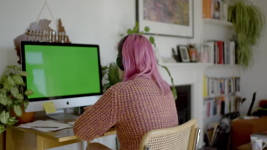 Woman with pink hair talks as she looks at a computer monitor