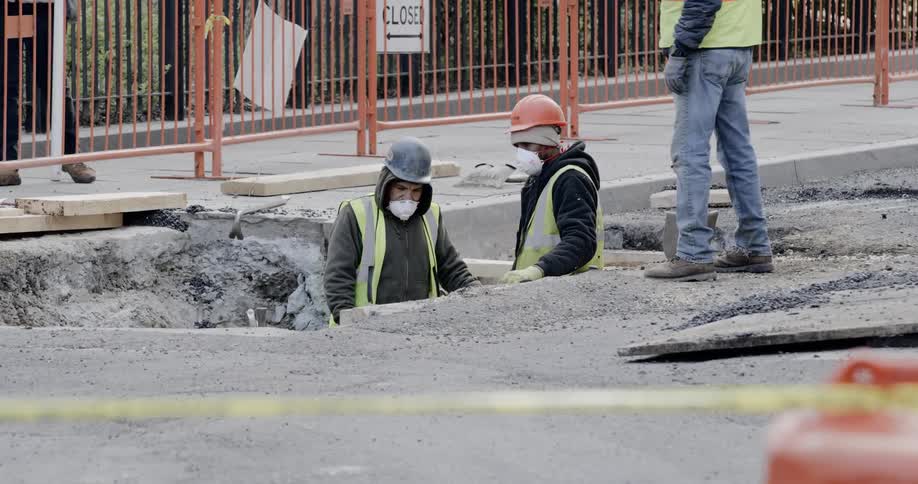 A construction worker carrying a shovel walks toward two other construction workers that are standing in a hole in the street