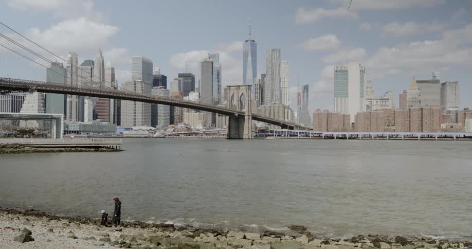 An adult and toddler walk away from a river coastline across from a large city