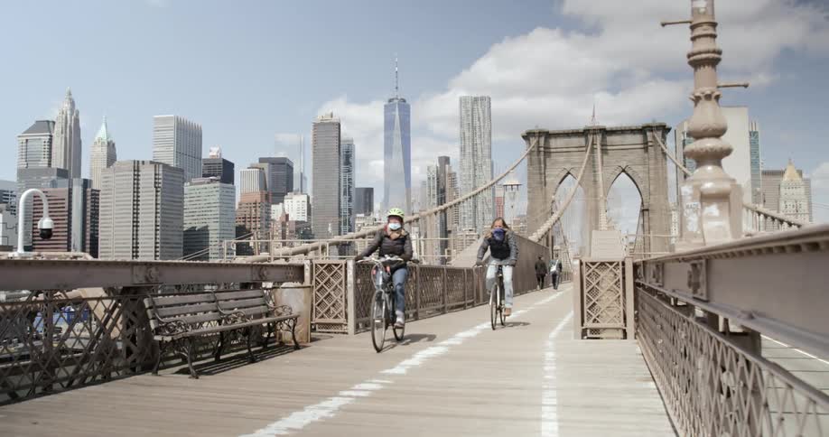 Bicyclists wearing face masks ride down a pedestrian bridge in front of a city
