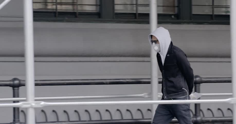 A man wearing a hoodie and a face mask strolls down an inner city sidewalk