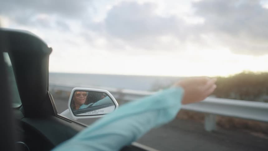 A woman sticks her arm out a car window as she drives by the ocean