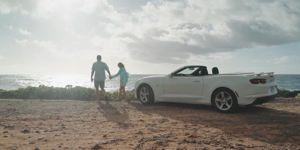 A man and woman get out of a convertible and hold hands as they walk to look at the water