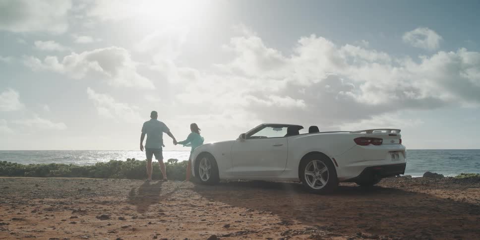 A man and a woman closing the doors of a car then walking and holding hands near the coast