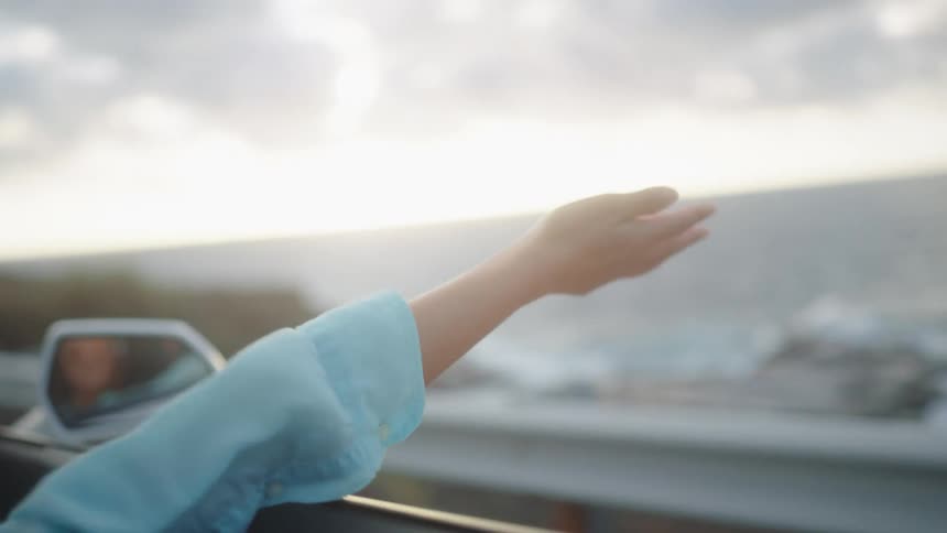 Woman extends arm out of convertible in breeze with coast in background