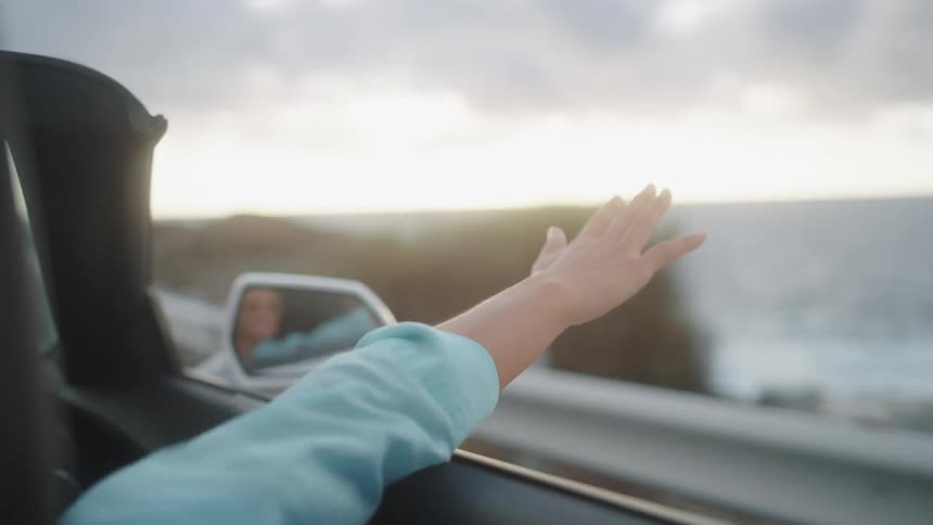 Young woman holds arm out of convertible in breeze while in car driving by ocean