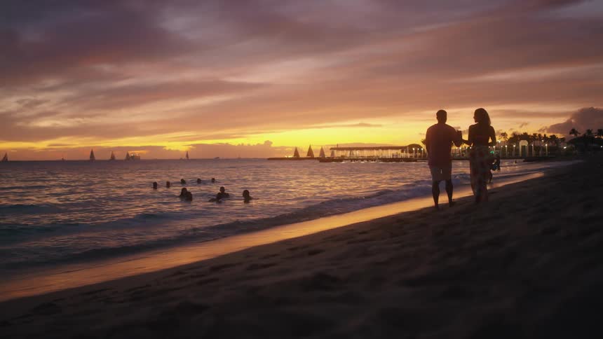 A man and woman walk down the beach holding hands while people swim in the ocean in the background