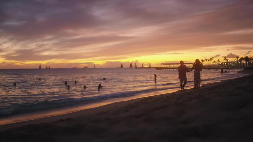 A man and a woman holding hands walking on the beach while people are in the water