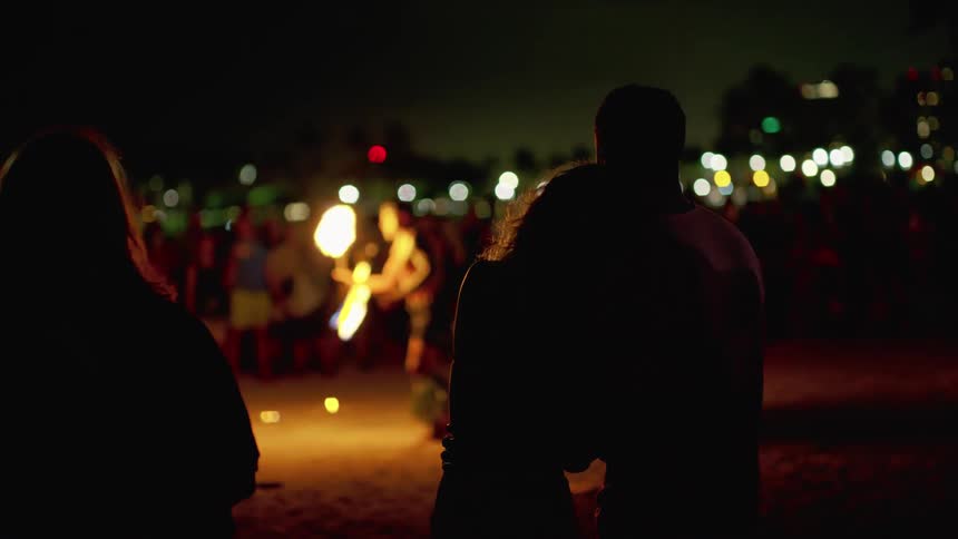 A woman with her head on her husbands shoulder as they stand with a crowd watching a hawaiian fire dancer at night