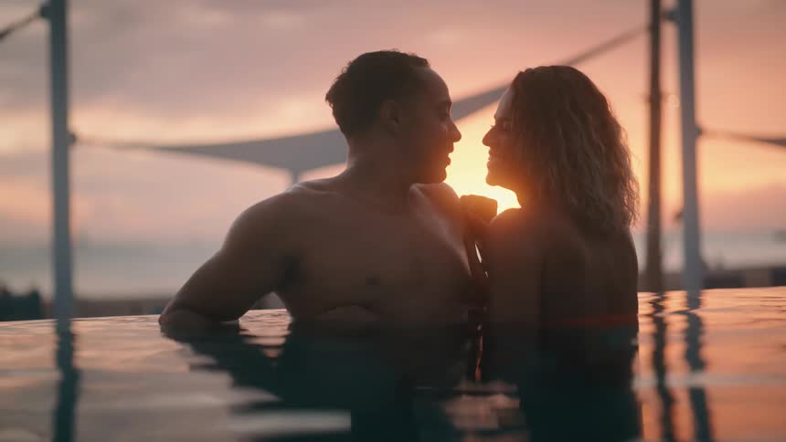 Man and woman flirt while standing in pool as sun sets behind them