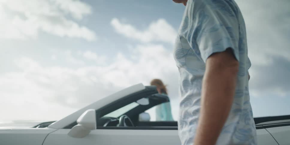 A man and woman get out of a convertible and hold hands as they walk towards the water